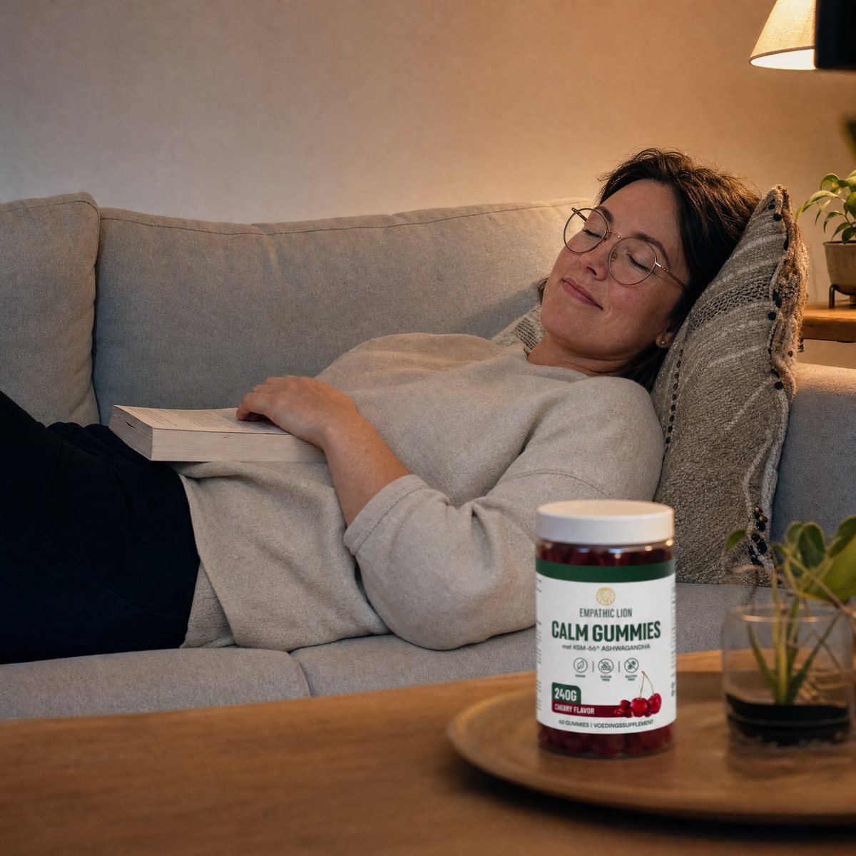 Vrouw ligt ontspannen op de bank met een boek, met een pot Calm Gummies op tafel